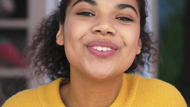 Close up view of Joyful african attractive woman in yellow sweater sending air kiss at the camera in living room