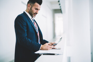 Side view of confident pensive proud ceo 50 years old reading incoming notifacation on smartphone device standing in office.Concentrated executive manager checking mail on mobile phone via 4G internet