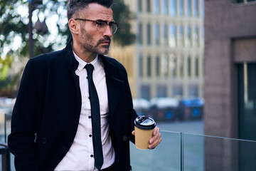 Successful mature business man dressed in trendy black coat holding urban coffee in hand.Concentrated beautiful economist in stylish eye glasses standing in city near office building during work break