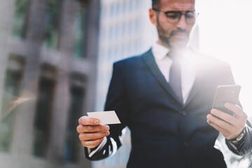 Selective focus on prosperous businessman holding visit card with mock up blank space calling taxi standing on blurred urban setting background, male entrepreneur dialing number on modern smartphone