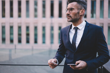 Handsome matured male entrepreneur in elegant suit standing outdoors on promotional background having informal meeting with colleague just contact him using information from visit card and smartphone