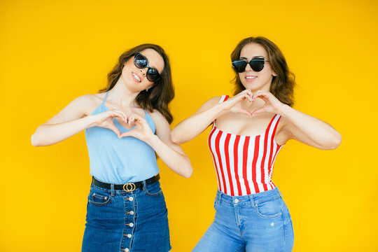 Image of two fashion women wearing girlish clothes posing at camera in stylish sunglasses isolated over yellow background