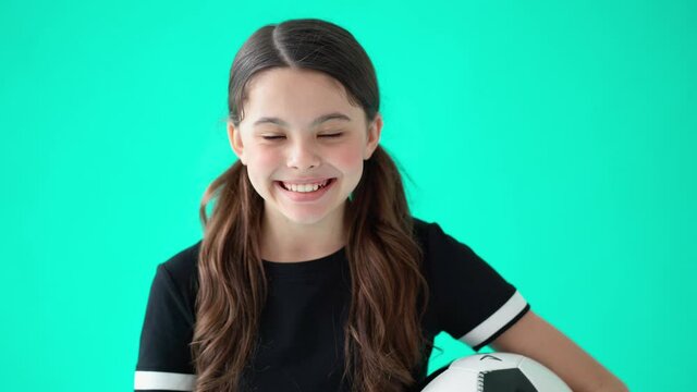 Head shot portrait of a cute teenage girl holding soccer ball, looking at camera and laughing, standing against turquoise blue background