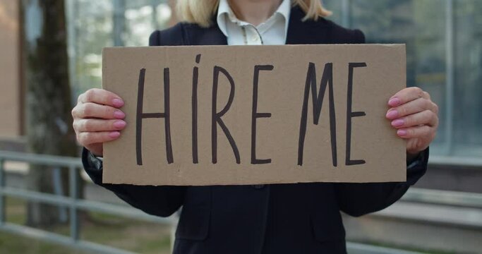 Crop view of female worker showing carton banner with hire me writing. Woman searching for job while standing at street. Concept of financial global crisis and unemployment