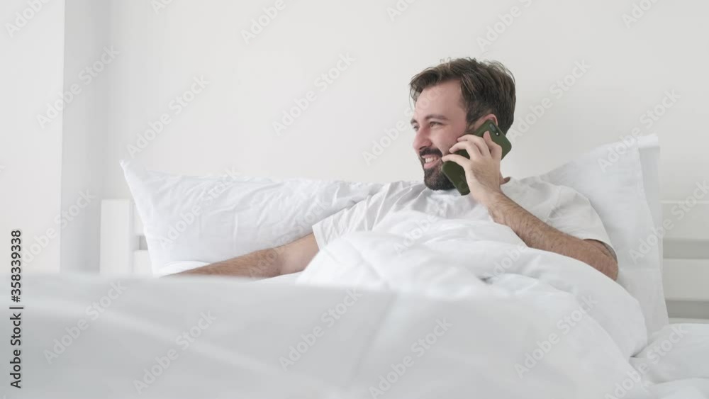 Sticker Handsome young happy cheerful bearded man indoors at home lies in bed while talking by cell phone