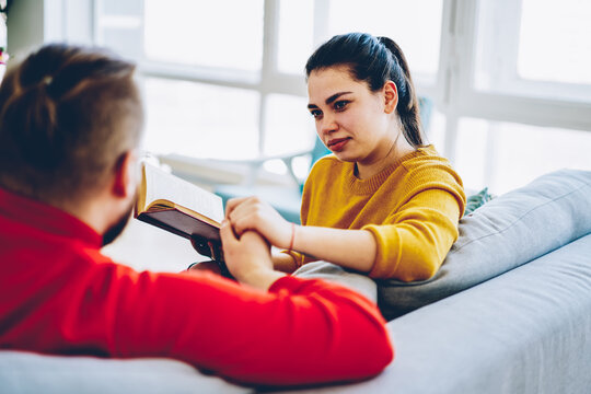 Pretty Woman Looking At Her Husband With Love Spending Free Time Together At Comfortable Apartment, Back View Of Hipster Guy Holding Hand Of His Girlfriend While She Sitting With Book At Home.