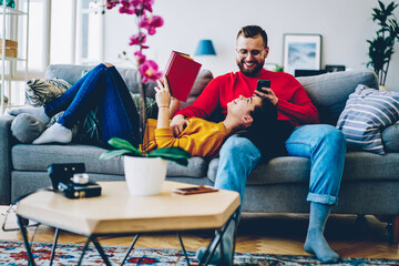 Happy young marriage communicating and laughing on free time at home interior , romantic couple on cozy sofa smiling and joking using smartphone for networking, woman reading book with boyfriend.