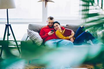Cheerful romantic couple browse website on smartphone read news together on free time, smiling woman watching video on mobile phone of her boyfriend lying on comfortable couch at living room .