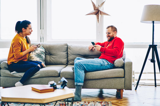 Happy hipster woman laughing and talking with her boyfriend sending her funny photos via smartphone, young romantic couple enjoying free time together share video using wireless internet at home.