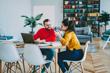 Fototapeta premium Positive couple using technology and wireless connection at home interior for freelance, woman using mobile phone for making booking online talking with her positive boyfriend working on laptop