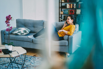 Naklejka premium Cheerful caucasian woman enjoying long mobile phone conversation with friend during free time at home satisfied with tariffs, happy hipster girl get good news during telephone talk sitting on sofa.