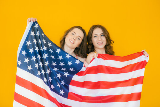 Image of two young beautiful girls wearing plaid shirts smiling and american flag together isolated over yellow background
