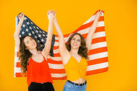 Image of two young beautiful girls wearing plaid shirts smiling and american flag together isolated over yellow background