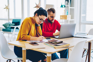 Obraz premium Young male and female sitting at desktop making research on netbook and writing information, serious woman in eyewear making accountings of bills and receipts for family budget man banking