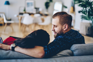 Romantic couple spending free time together at home interior talking to each other, back view of hipster girl lying on her boyfriend hand relaxing during communication at cozy couch in apartment.