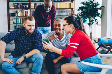Cheerful group of male and female friends watching online video on smartphone sitting at couch at living room, hipster girl shoving photos on mobile phone to multiracial colleagues  having fun.