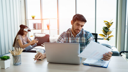 Young caucasian man use laptop at home or modern office his wife playing virtual reality(VR) in the background with copy space.work from home concept.Quarantine due to coronavirus.