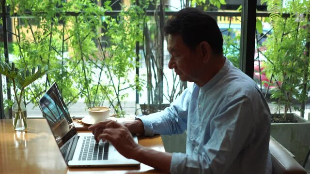 Old man using laptop computer in cafe and wearing mask for prevent the spread of the Covid-19, Covid-19 virus protection concept in Thailand.