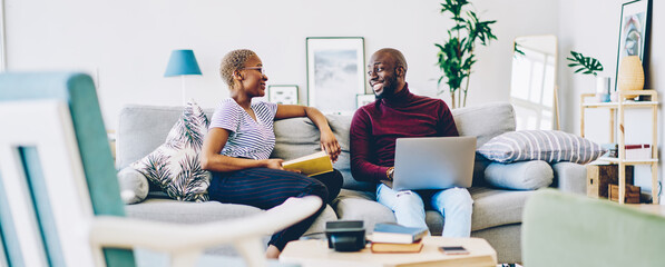 Cheerful african american marriage spending time at home enjoying free time together, positive woman sitting with book while communicating with her husband using laptop computer and wifi in apartment.