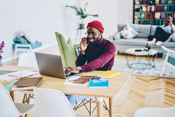 Happy dark skinned male satisfied with mobile phone conversation with customer support installing app on laptop computer,smiling african american man freelancer satisfied with online business.