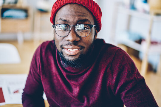 Close up image of dark skinned hipster guy in trendy hat and eyewear for vision protection looking at camera, portrait of smiling bearded male satisfied with stylish spectacles sitting indoors