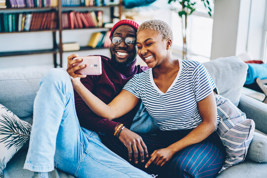 Cheerful dark skinned couple in love resting at home interior posing for selfie on smartphone camera,happy young hipsters making image on modern mobile phone laughing at living room together.