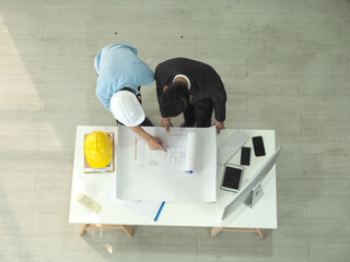Top view of asian businessman and architect or engineer are discussing work together in office.Man pointing at blueprint.Foreman is wearing construction helmet.