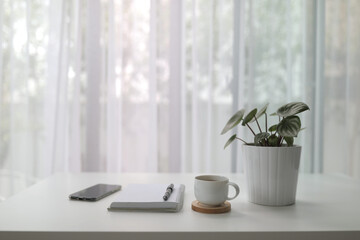 coffee cup with plant and notebook pen and phone on white desk with see through curtain 