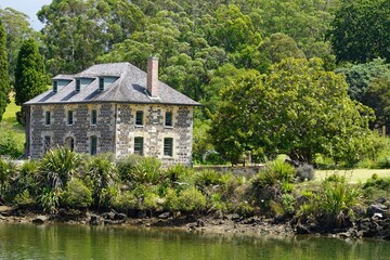 Fototapeta na wymiar Historical Mission Station - Stone Store house in Kerikeri surrounded by green trees and plants. Lake and shore on front.