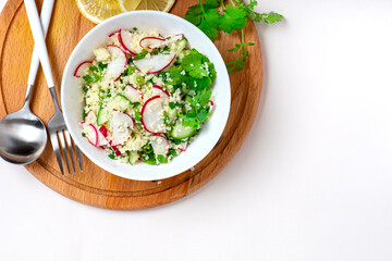 Fresh summer vegetable salad of couscous, radish, cucumbers, parsley and lemon in a bowl on a light background, top view, copy space. Vegan food. Spring or summer food. Free space for text.
