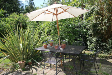Landscape of Summer garden with outdoor seating table and metal chairs with giant parasol wood on stone patio outdoors and exotic tropical plants, bamboo and hedging, grass lawn, plants in background © Carmina