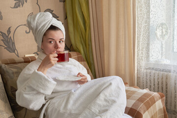 A girl in a white bathrobe is relaxing in a spa on the couch drinking tea.