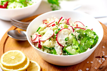 Fresh summer vegetable salad of couscous, radish, cucumbers, parsley and lemon in a white bowl on a served table. Vegan food. Spring or summer food. Salad with couscous and raw vegetables close-up.