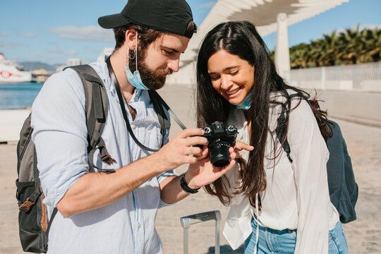 Couple With A Mask Pulled Down Over Their Chin Looking At A Camera With A Suitcase And A Map In Hand