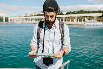 Man with a cap and a digital camera hanging from his neck looking at a map in front of the sea