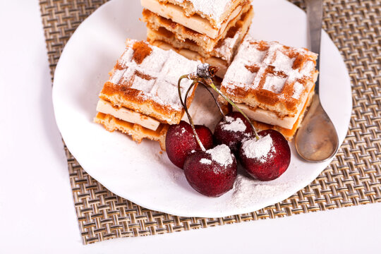 Belgian waffles in powdered sugar with bananas and cherries on a white plate. In the background, a teapot and a Cup for morning tea. Delicious 
