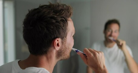 Brushing teeth man looking in mirror of home bathroom using toothbrush in morning routine for clean dental oral care. 4K (UHD) 59.94 FPS SLOW MOTION.