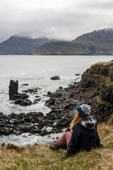 Fototapeta premium A beautiful girl with long blonde hair and in warm clothes is sitting on the cliffed coast of the Atlantic Ocean. She admires the Icelandic landscape on the background of a volcanic beach