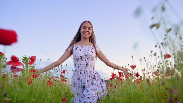 Beautiful smiling teen girl in white dress run and having fun through a poppy field. Happy child playing on meadow of blooming red poppy at sunset. Leisure in nature.