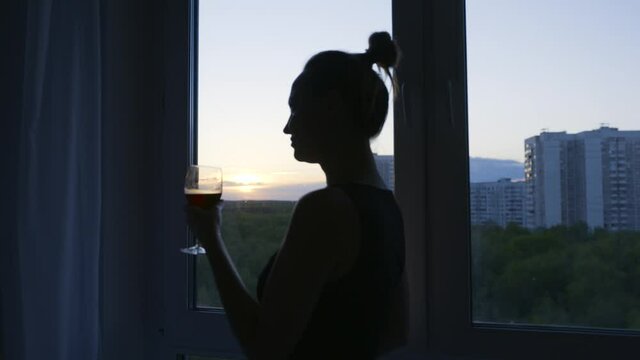 Woman drinks wine with pleasure on background of window. Action. Attractive woman drinks of red wine at home. Silhouette of woman drinking wine on background of evening window