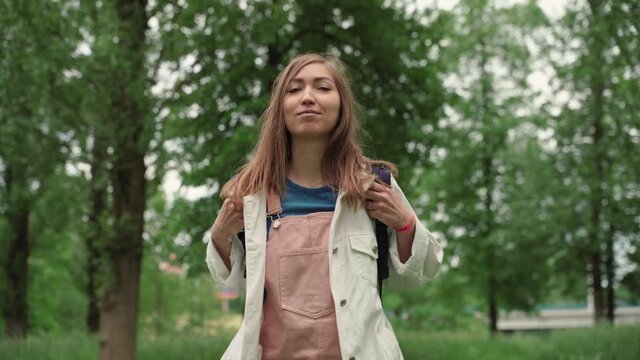 Beautiful woman smiles walking along path surrounded by foliage and trees. Front view of female with backpack walking at the summer park.