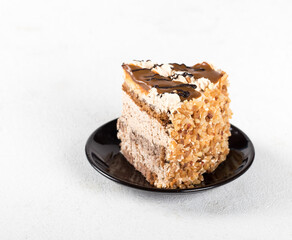 A piece of sponge cake with cream, caramel, nuts and chocolate on a plate on a light background
