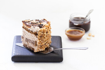 A piece of biscuit cake with cream, caramel, nuts and chocolate on a wooden Board on a light background