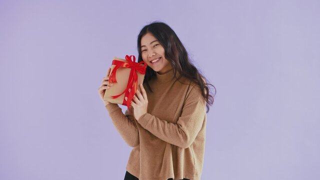Cute asian model in brown sweater. She is smiling and holding gift box tied with red ribbon while posing against purple studio background. Close up