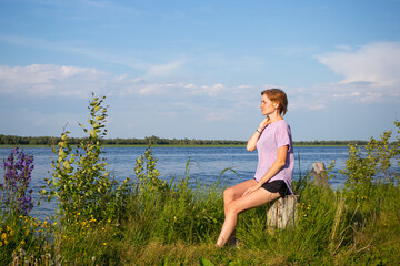 Fototapeta premium The concept of walks alone. Young girl with short hair in purple T-shirt and shorts sits on river bank. Summer sunny evening. Copyspace. Cozy cute rural landscape..