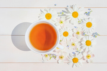 Chamomile tea in a white little cup on a light wooden surface. Around the cup are scattered flowers. The concept of natural vitamin drinks for health, immunity. Top view, flat lay..