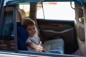 Happy caucasian boy sitting in a blue child safety seat and showing thumb. Traveling with a child in a car with a leather interior. Portrait of a cute preschooler wearing a seat belt and ready to ride