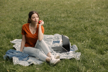 Full length portrait of dreamy girl eating apple while sitting on the green grass in the city park with copy space. Pretty woman have a break after distance work. 