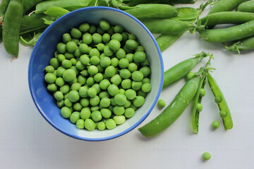Fresh peeled green peas in a bowl and open pods on a white wooden table background with copy space. Top view