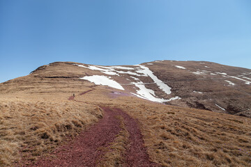 Distant mountain hiker hiking towards distant mountain peak on a curvy mountain track covered by remaining snow at spring and clear, blue sky